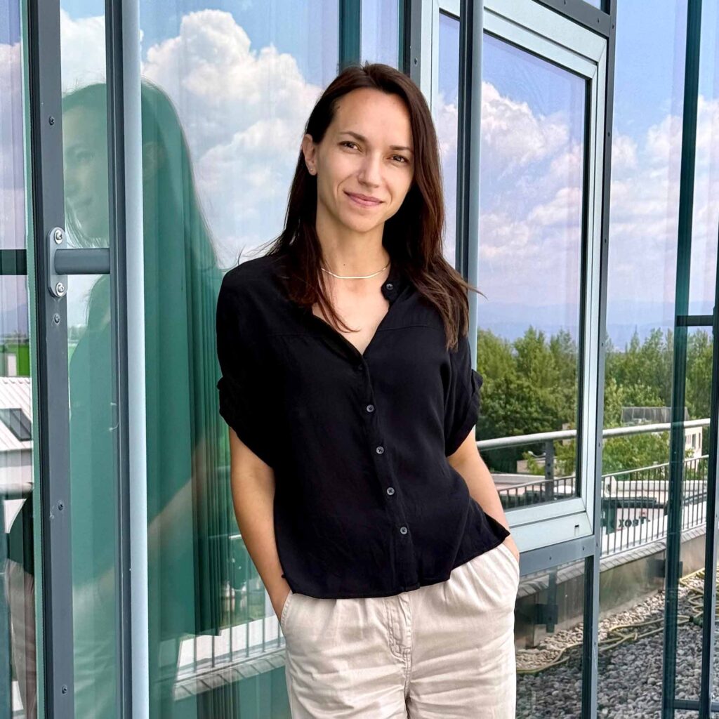 Eine Frau mit dunkelbraunem Haar und schwarzer Bluse steht vor einer Glasfassade und lächelt in die Kamera.
