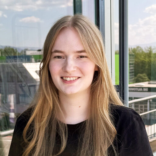 Eine Frau mit dunkelblonden Haaren und einem schwarzen T-Shirt steht vor einer Glasfassade und lächelt.