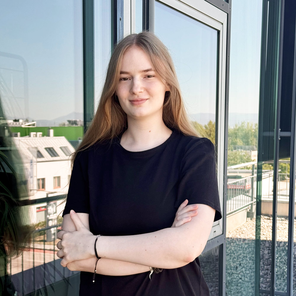 Eine Frau mit dunkelblonden Haaren und einem schwarzen T-Shirt steht vor einer Glasfassade mit verschränkten Armen.